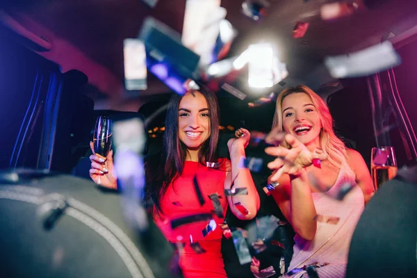 Beautiful people are sitting in car and parting. They are throwing confetti on camera. Young women are holding glasses of champagne. They are very happy and excited. — Stock Photo, Image