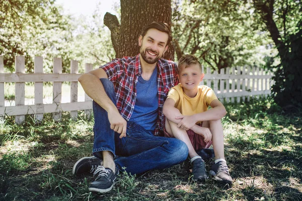 Pěkný obrázek otce a syna seděli pod stromem a při pohledu na fotoaparát. Oni se usmívají. Dospělý je objímání dítě. — Stock fotografie