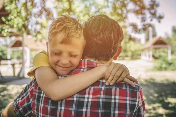 Un niño alegre y feliz está abrazando a su padre. El chico mantiene los ojos cerrados y sonriendo. Están de pie afuera en el parque . — Foto de Stock