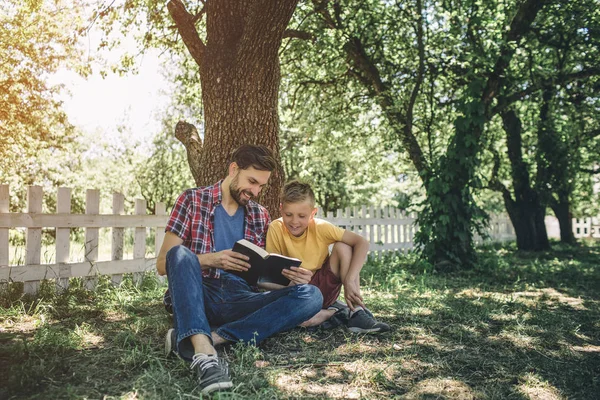 Cuidado un buen padre está sentado con su hijo inder árbol y la celebración de libro. Lo están mirando. Lo están leyendo. El chico está muy interesado. Parece feliz. . — Foto de Stock