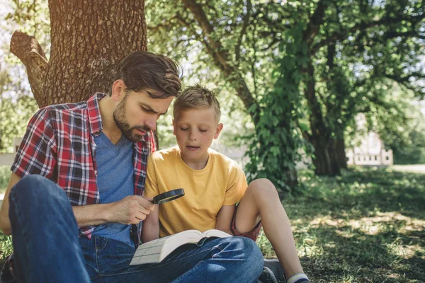 Gente seria y concentrada están sentados juntos bajo el árbol y mirando el libro. El adulto lo sostiene. También está mirando el libro a través de Lenz. El chico también está mirando el libro. . — Foto de Stock