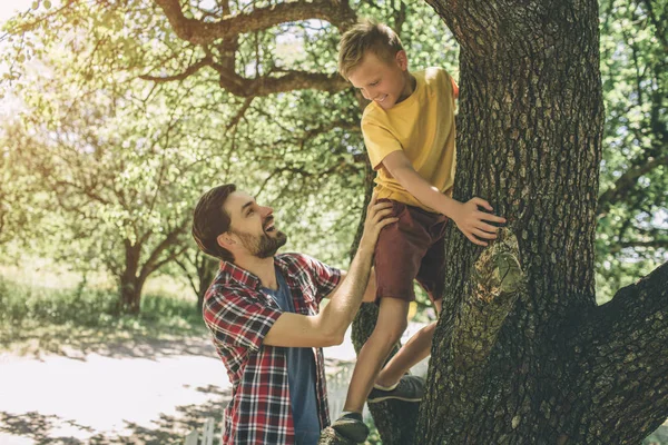 El barbudo está parado cerca de un árbol y apoyando a su hijo. El chico está subiendo. El niño ha puesto sus manos en el árbol y mirando a su padre. Están sonriendo. . — Foto de Stock
