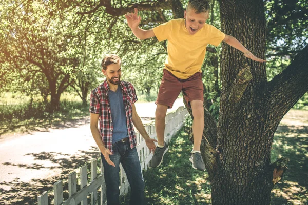 Image émotionnelle d'un enfant heureux sautant sur le sol. Il fait ça à partir d'un arbre. Père regarde ça et sourit aussi . — Photo