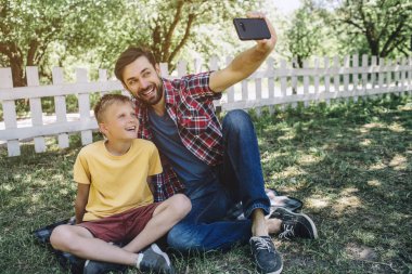 Adam onun benzeri çim oturan ve telefon elinde tutarak. O çocukla selfie alıyor. Bunlar miling ve kameraya poz.
