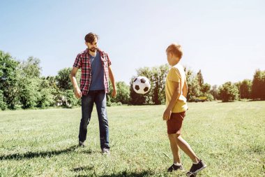 Baba nd oğul futbol oyunu oynarken bir resim. Babamdan oğlu için uçan bir top var. Bu havada. Sahada dışında güneşli.