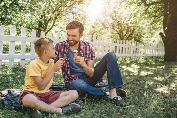 Otec a syn sedí společně na dece a pohledu na sebe. Oni se usmívají. Zmrzliny mají v rukou. Chlapci se těší společnosti navzájem. — Stock fotografie