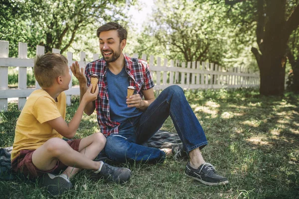 Feliz padre está sentado junto a su hijo en manta y sonriéndole. Tanto él como su hijo comen helados. Los chicos están disfrutando pasar tiempo juntos . — Foto de Stock
