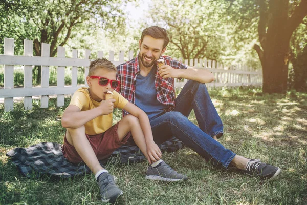 Père est assis avec son fils sur une couverture. Il regarde l'enfant et sourit. Le gamin porte des lunettes et mange de la glace. Il a l'air cool . — Photo