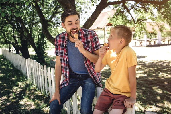 Šťastný a nadšený, rodič je při pohledu na svého syna a jíst zmrzlinu. Dítě s vážný pohled při pohledu na otce a jíst i zmrzlinu. Sedí na plotě. — Stock fotografie