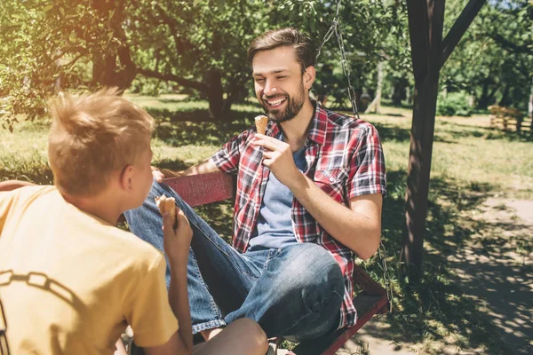 Una imagen de un hombre barbudo feliz sentado sobre una manta con las piernas cruzadas y mirando a su hijo. El tipo tiene helado en las manos y sonríe. El niño también come helado. . — Foto de Stock