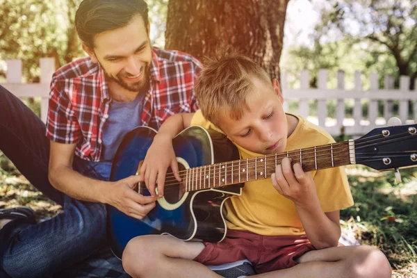 Talented boy is looking on strings and playing guitar. His father is sitting besides him and helping him. Adult is smiling.