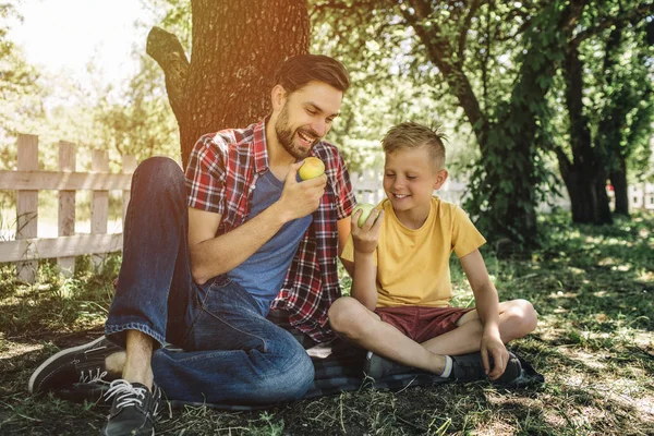 El hombre está sentado sobre una manta con su hijo. El tipo está mirando a los niños manzana y sonriendo. El chico está mirando allí también. Se está preguntando . — Foto de Stock