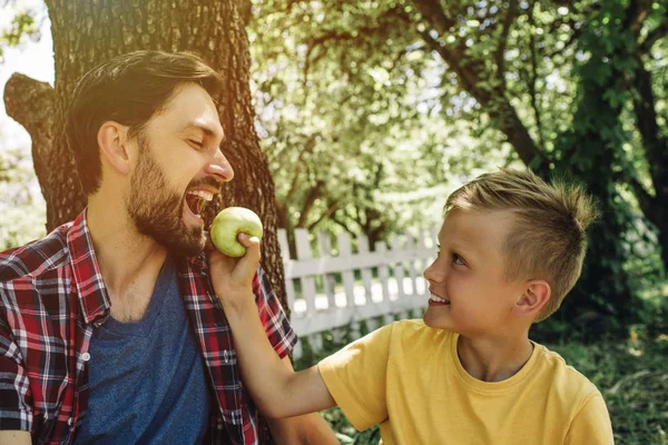 Linda imagen de padre e hijo sentados juntos fuera bajo el árbol. Chico está sosteniendo manzana mientras su padre está mordiendo pedazo de ella . — Foto de Stock