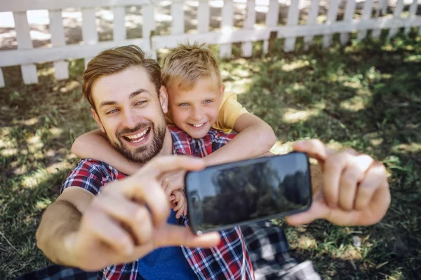 Adult and kid are looking on phone and smiling. Guy is holding device while child is hugging his dad. This picture is cute and awesome. — Stock Photo, Image