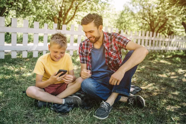 Malý chlapec sedí na trávě se svým otcem se jejich nohy překřížené. Dítě drží telefon v ruce a při pohledu na něj. Usmívá se. Táta se dívá na zařízení příliš a usmívající se také. — Stock fotografie