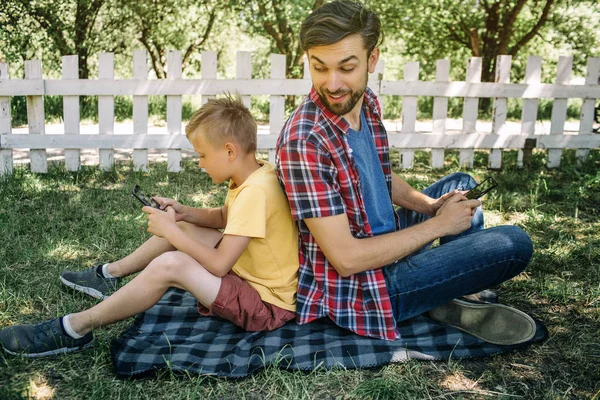 Consentrated chico está sentado espalda con espalda con su padre y jugando juegos en el teléfono. Papá está mirando a su hijo y sonriendo. Él también tiene un teléfono. . — Foto de Stock