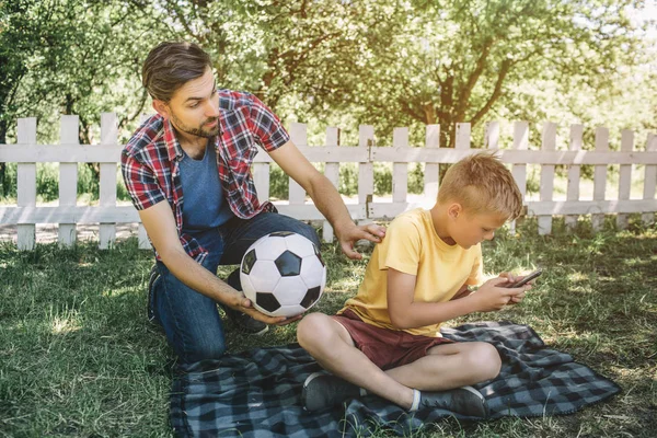 Dobrý otec se při pohledu na svého syna a drží míč. Chce hrát se svým synem. Dítě si hraje na telefonu. On nechce hrát fotbal. — Stock fotografie