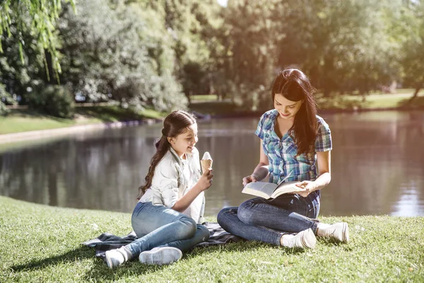 Smart woman is sitting at the edge of small lake with her daughter and reading a book. Her kid is sitting besides her and eating ice cream. Girl is looking at what mom is doing right now