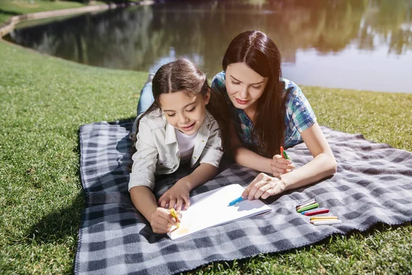 Las niñas están tumbadas en la manta este hermoso día soleado en la hierba. Están usando colores azul y amarillo para dibujar. Las niñas se ven veri interesado . — Foto de Stock