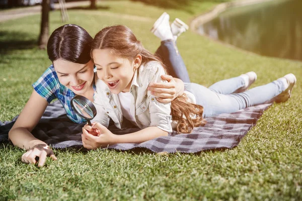 Interested and smart girl is lying on blanket with her mom and holding loop in hands. She is wondering. Her mom is embracing her daughter and pointing on grass. They are smiling.