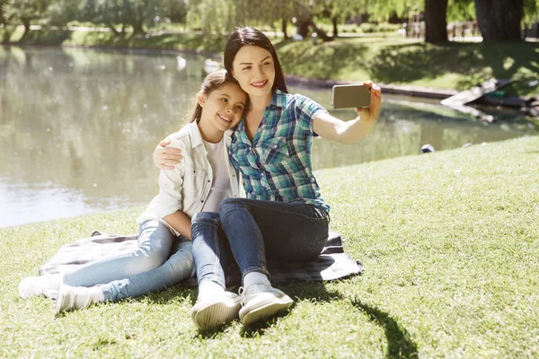 엄마와 딸 복용 selfie의 좋은 그림. 그들은 담요 연못 근처에 앉아 있다. 여자는 전화를 들고 있다. 그들은 그것을 보고 하 고 웃 고 있다. 여자는 포즈. — 스톡 사진