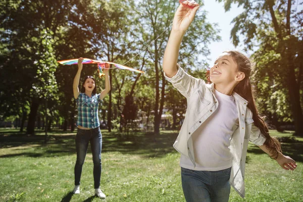 Kvinnan är stående bakom flicka och hålla kite. Flickan är att dra tråd av kite framåt. Hon tittar på kite och leende. — Stockfoto