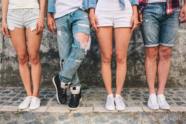 Uma foto de pernas de adolescentes. Três deles estão em pé direito enquanto o homem de jeans está segurando a perna esquerda sobre a direita. As raparigas usam calções e um rapaz também. Eles estão de pé sobre fundo cinza . — Fotografia de Stock