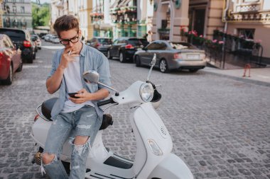 Yakışıklı olan motosiklet üzerinde oturan ve telefon elinde tutarak. O bakıyor. Theere diğer elinde bir sigaradır. O sigara içiyor. Adam bekliyor.