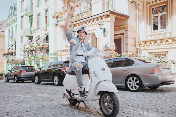 Man in casual clothes is riding on motorcycle. He is wearing helmet on head. Guy is holding fist in air and looking to the side. He is riding on road.