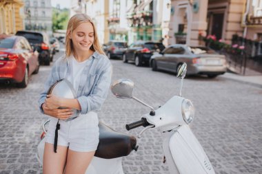 Pozitif ve attractiwe genç kadın motorcyles kenarında oturan aşağı bakıyor nad koltuk. Gülümsüyor. Kız kask iki eliyle tutuyor.
