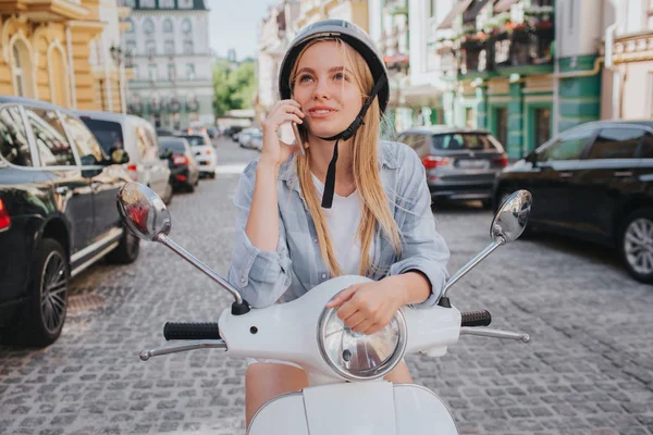 Menina loira lindo no capacete está posando na câmera. Ela está sorrindo. A rapariga está sentada numa mota no meio da rua. Ela parece feliz. . — Fotografia de Stock