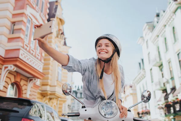Lány ül, és támaszkodva a motorkerékpárok vezérlő headling és a selfie figyelembe véve. Kalliopé telefon a kezében. Lány keres, a kamera, és mosolyogva. — Stock Fotó