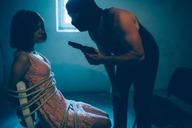 Maskeli adam kızın önünde duran ve ona bakıyorsun. Silah düzenliyor. Kız kaçırdı. Adama bakıyor. Kız ile ipleri sandalyeye bağlı.