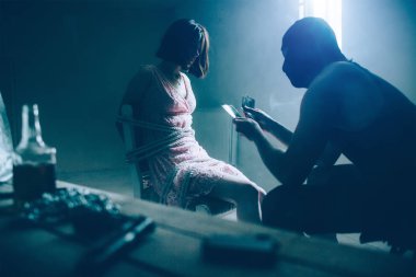 Katil kurbanı önünde oturan ve faturaları para sayma. Esmer bağlı elleriyle sandalyeye oturmuş ve bir adam arıyorum. Maske takıyor. Onlar tablo oturuyorlar bootle, nerede silah.