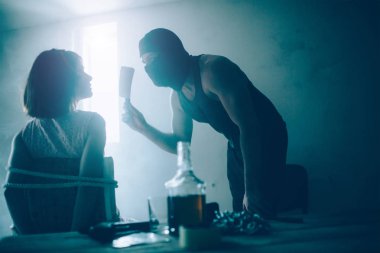 Maskeli adam kaçıran bağlı kızın önünde duran ve kalın bıçak elinde tutuyordu. O kurbana arıyor ve onu korkutmak istiyor. Kız ona bakıyor. Alcoholm masada şişe vardır.