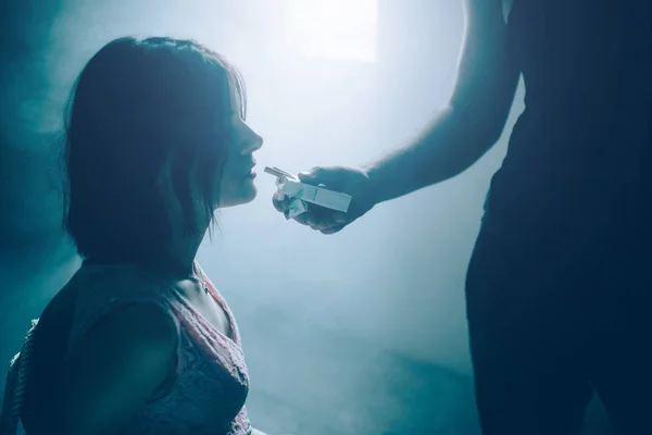 Ortrait de menina sentada amarrada e olhando para pequena caixa de cigarros que cara em camisa preta está segurando na frente dela. Ela não quer fumá-lo. Estão perto da janela brilhante . — Fotografia de Stock