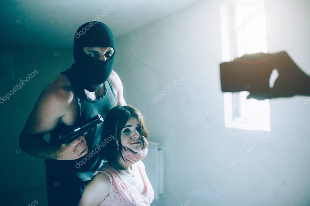 Angry kidnapper is holding girls head with lefy hand and gun in another one. He is looking on camera with angry eyes. He wears mask. Girl is looking at phone as well. She is terrified and afraid.
