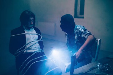 Uzun ve karanlık saç s onun kaçıran önünde oturan adam. Halatlarla bağlanmış. Ağzını musluk ile kaplıdır. Maskeli katil işadamı seyir ve ışıkları ve silah elinde tutan.