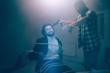 Adam kaçırma video kaydediyor. Manyak ayakta ve elinde silah kurban yakın baş ve işaret kamerada. Sakallı adam bakmak ve ağlıyor. O acı ve halatlar ile bağlı..