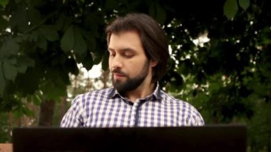 Yakışıklı adam ağaçların altında oturan ve dizüstü tutarak. O ekrana bakarak ve çalışma. Adam Kupası ve bunu alıyor. Ekrana bakar ve gülümsemeye başlar. Adam içki Kupası tekrar.
