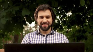 Adam ağaçların altında oturan ve dizüstü tutarak. Gülümse ve fincan kahve ve içki almak başlar. Adam ciddi görünüyor. O aşağı ve tekrar ekran ve içki froom Kupası için arıyor ve geri koymak.