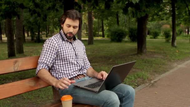 Brodaty mężczyzna siedzi na ławce w parku i pracy z laprop. On jest wpisywanie i biorąc filiżankę kawy. Człowiek jest przesuwanie palcem na touchpad i picie z kubka. Stawia go z powrotem. na ławce. — Wideo stockowe