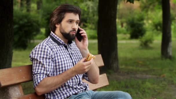 El hombre de negocios está sentado en Bech en Park y hablando por teléfono. Muerde un pedazo de hamburguesa y sigue hablando. El hombre pone el teléfono en otra oreja y pone hamburguesa en la boca. Toma cuaderno y blanco abajo . — Vídeos de Stock