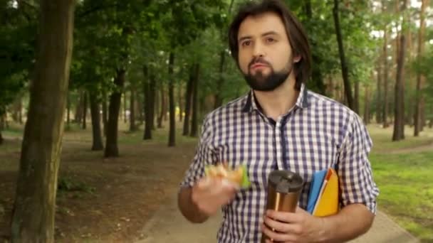 Mann läuft auf der Straße im Park und hält Tasse, Burger und Ordner in der Hand. Er schaut geradeaus. er kaut. dann schaut Mann auf Burger und beißt wieder in ein Stück. — Stockvideo