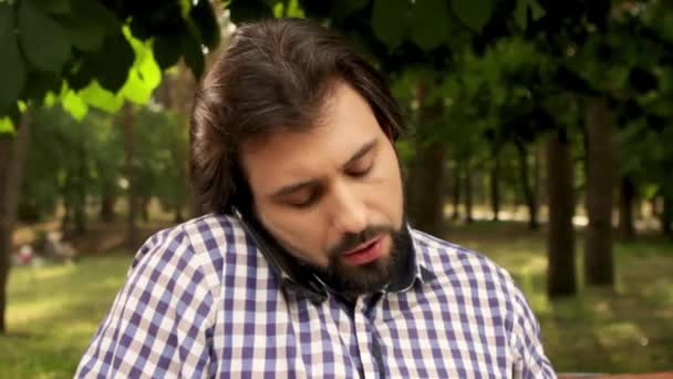 Close up of man sitting in park and talking on black phone. He changes position phone and looking down. Then guy put off black phone and take white one and starts to talk. And he changes phones again. — Stock Video