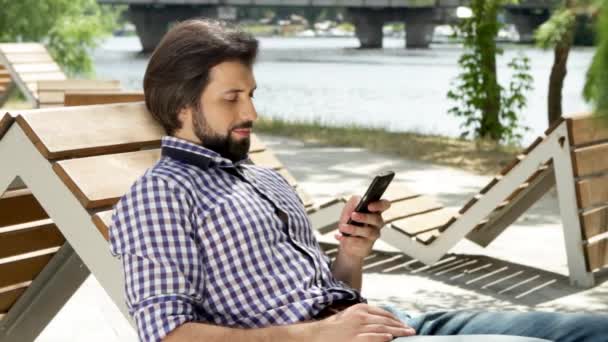 Hombre Está Tumbado Banco Sosteniendo Teléfono Está Mirando Escribiendo Mensaje — Vídeos de Stock