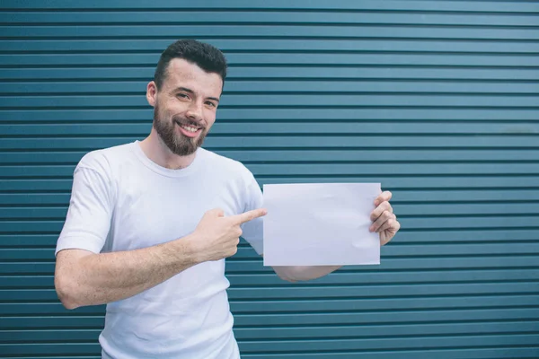 Positive man is standing and holding white piece of paper in left hand and pointing with finger on right one. Guy is looking on camer and smiling. Isolated on striped and blue background. — Stock Photo, Image