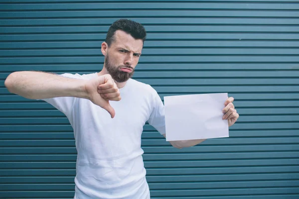 Unhappy man is standing and looking oon camera. He is posing. Guy is holding piece of paper in left hand. He is showing big thumb down. Isolated on striped and blue background. — Stock Photo, Image