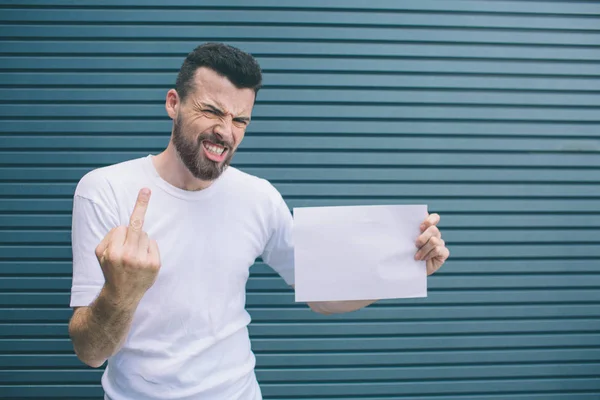 Angry man is standing and posing on camera. He is showing third finger. It is bad sign. Also guy is holding blank piece of paper. Isolated on striped and blue background. — Stock Photo, Image