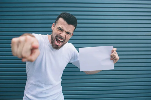 Guy is standing and posing. He is looking and pointing on camera. Also man is holding blank piece of paper in left hand. He is angry. Isolated on striped and blue background. — Stock Photo, Image